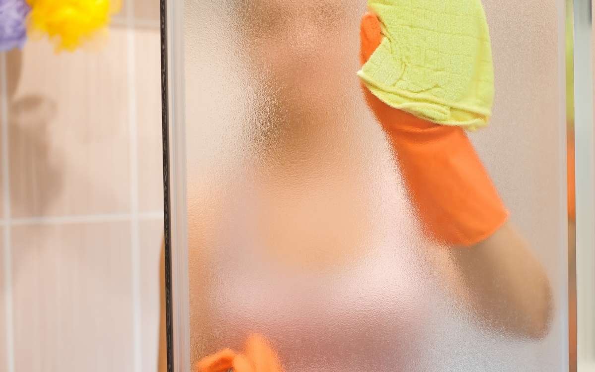 a person behind the glass shower door holding a yellow rug wiping the glass door