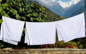 White bedsheets hanging to dry on clothes line with bricks underneath and plants at background