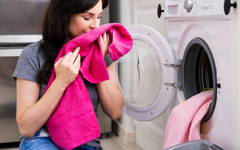 a woman smelling a pink-colored towel beside a washing machine