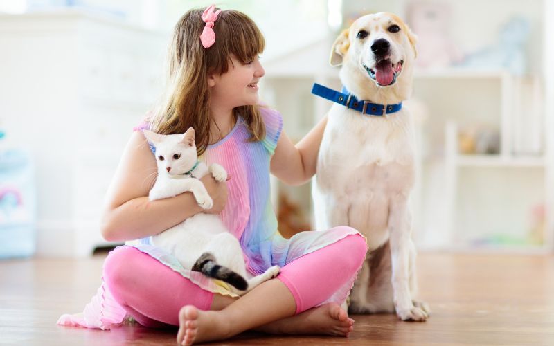 A girl cross-sitting on the floor while holding a white cat and a dog inside a clean house