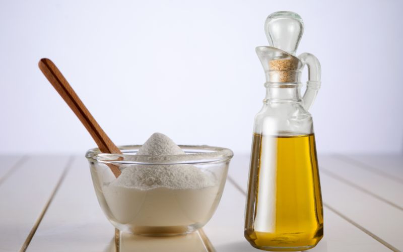 A glass bowl filled with white powder with a wooden spoon beside a bottle of oil