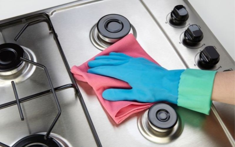 A hand with blue gloves uses a pink rug for cleaning the stove top