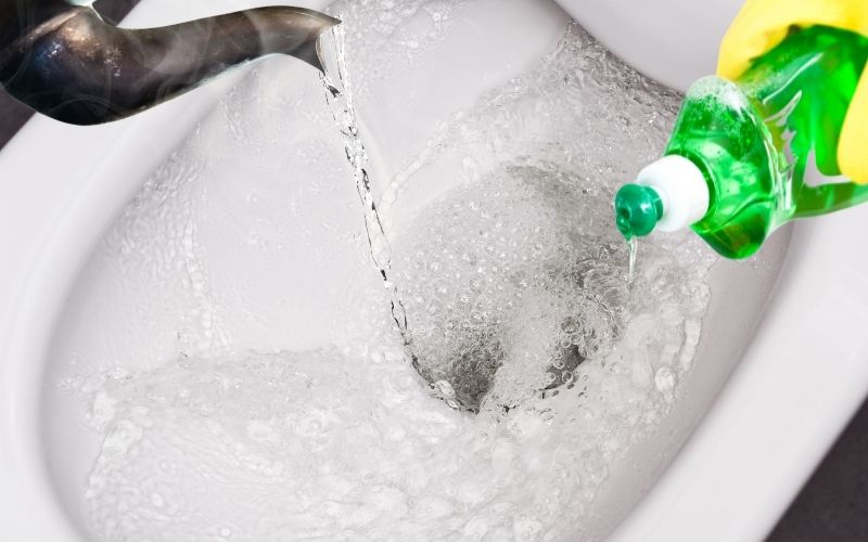Hot water and dishwashing soap pouring ove a toilet bowl to help unclog it.