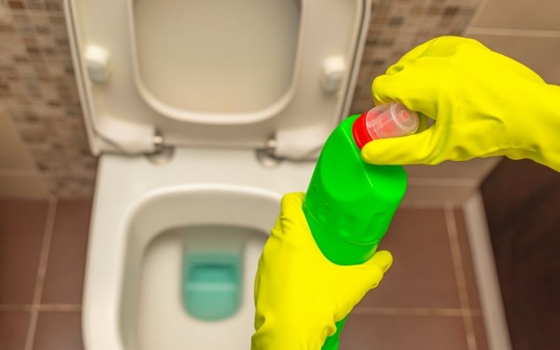 Photo showing two gloved hands holding a drain cleaner in green bottle over a toilet.