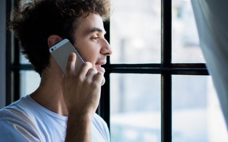 Photo of a man holding a phone to his ears beside a window