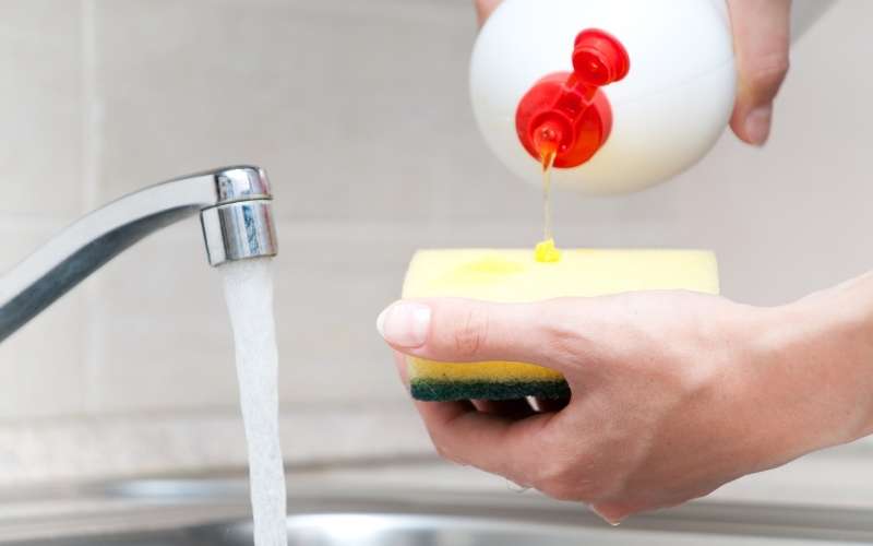 Photo of a hand holding a sponge, a bottle of dishwashing soap and a faucet with flowing water
