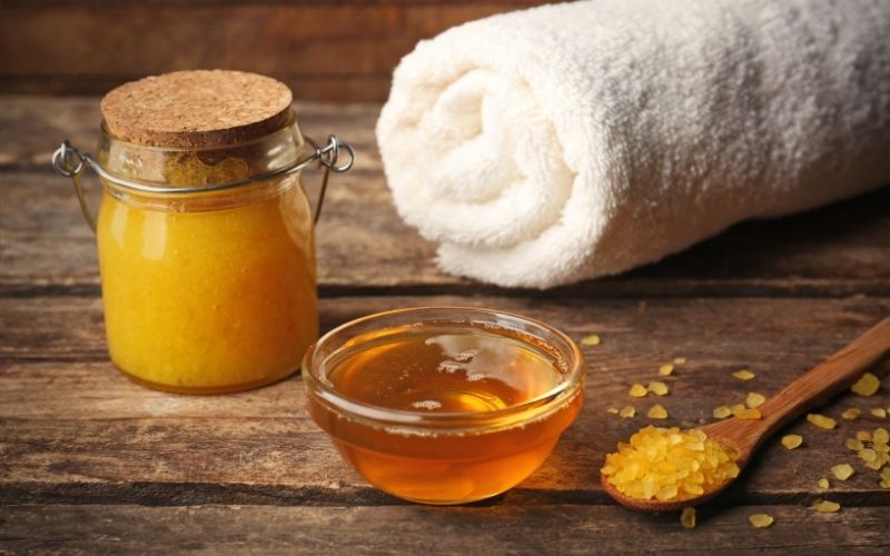 A natural scrub in a small jar, a bowl of honey a spoon of salt and a roll of white towel that can use as body wash