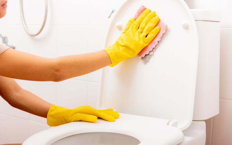 A pair of hands with yellow gloves cleaning the toilet seat using a cloth