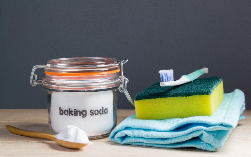 Photo showing a jar of baking soda, a wooden spoon with white powder, clean rag, a tooth brush and sponge placed on a table top